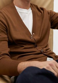 Cardigan marrone realizzato in un materiale morbido con scollatura a V, due bottoni e polsini a coste, indossato sopra una t-shirt bianca.