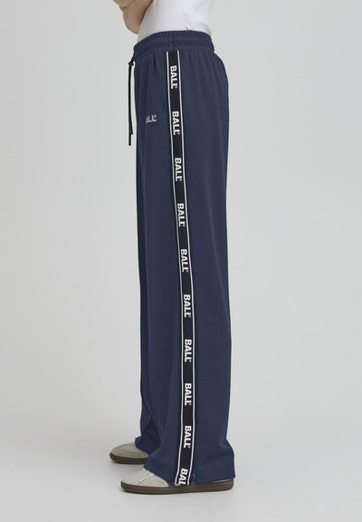 Pantalon de survêtement bleu marine avec une taille élastique, présentant des rayures blanches sur les côtés avec "BALL" répété. Le matériau semble doux et confortable.