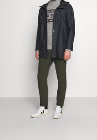 Waterdichte donkerblauwe jas met een capuchon, grijze kabeltrui, olijfgroene broek en witte sneakers met zwarte accenten.