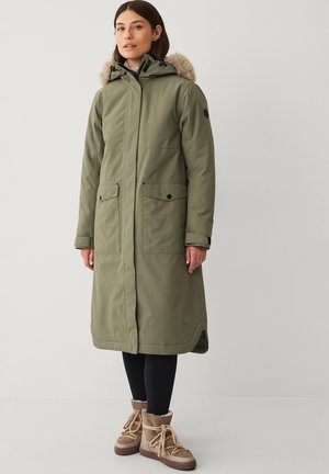 Oliven grøn parka med hætte foret med pels, lynlås foran, to store frontlommer og ribbet manchetter. Båret med sorte leggings og brune støvler.