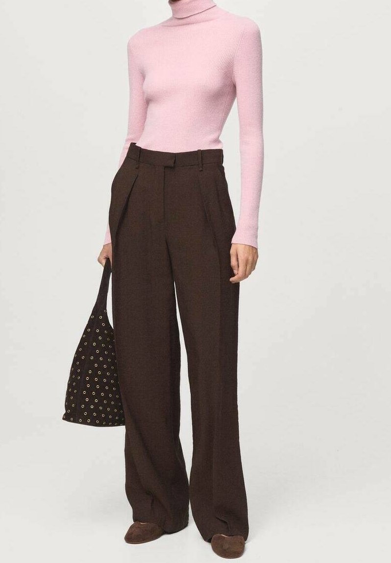 Personne portant un pull à col roulé rose et un pantalon marron taille haute à jambes larges, tenant un sac cabas noir clouté.