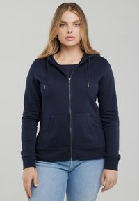 Kobieta o prostych blond włosach, ubrana w granatową bluzę na zamek z przednimi kieszeniami oraz jasnoniebieskie dżinsy, stojąca na jednolitym tle.