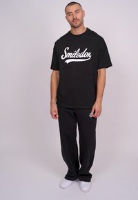 Zwarte oversized t-shirt met wit "Smilodox" logo, gecombineerd met zwarte broek. Casual ontwerp, zachte stof en een relaxte pasvorm. Witte sneakers inbegrepen.