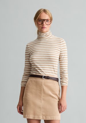 Jonge vrouw met een grote bril, beige-wit gestreepte coltrui, beige corduroy rok en een bruine riem tegen een effen grijze achtergrond.