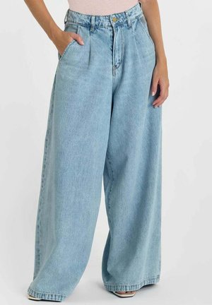 Vrouw die lichtblauwe denim jeans met hoge taille, wijde pijpen, voorplooien en zakken draagt, staand met één hand in de zak.