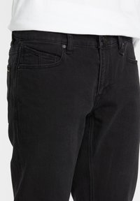 Jean en denim noir porté avec une chemise blanche, avec des poches avant et une poche montre, ainsi que des passants de ceinture à la taille.