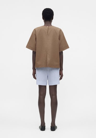 Άτομο που στέκεται με την πλάτη, φοράει χαλαρό καφέ μπλουζάκι με κοντά μανίκια, ανοιχτό μπλε σορτς και μαύρα παπούτσια χωρίς κορδόνια σε απλό φόντο.
