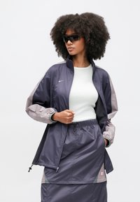 Ναυτικό αδιάβροχο με ανοιχτό γκρι μανίκια, ελαστικά μανσέτες και ψηλό γιακά. Συνδυάζεται με παντελόνια με κορδόνι στην ίδια απόχρωση.