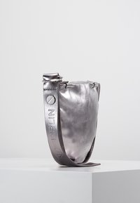 Metallinen hopeanvärinen nahkakäsilaukku, jossa on pehmeä, pyöreä muoto. Varustettu leveällä hihnalla, johon on painettu "BERLIN" ja pyöreällä metallikoristeella.