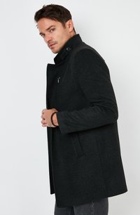 Abrigo de mezcla de lana negro con un cuello alto, bolsillos laterales y un bolsillo en el pecho con cremallera. Presenta detalles de botones en el cuello.