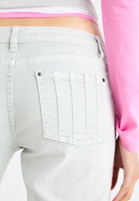 Jeans de mezclilla gris claro con un diseño de bolsillo trasero plano, con detalles de costura sutiles y un acento metálico. Parte superior de manga larga rosa brillante visible.