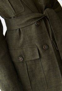 Olivenfarvet bæltemantel med struktureret stof, to forlommer og brune knapper med fire huller, der fremhæver en skræddersyet design.