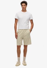 T-shirt bianco a girocollo, pantaloni corti beige e sneakers beige. Tessuto morbido, vestibilità rilassata, con un logo sottile sulla maglietta e calze con texture.