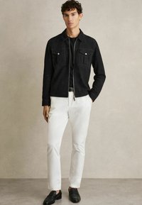 Veste en daim noir avec deux poches poitrine, pantalon blanc et mocassins noirs. Design simple, coupe près du corps et texture lisse.