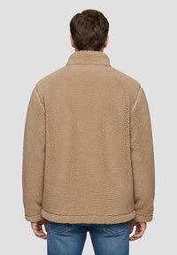 Giacca in pile marrone chiaro con superficie strutturata, colletto alto e vestibilità morbida, vista da dietro su sfondo grigio.