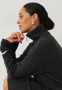 Mørkegrå højhalset sweater lavet af blødt, tekstureret materiale. Har lange ærmer, tætsiddende silhuet og subtile detaljer ved manchetterne.
