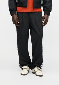 Άνδρας που φοράει μαύρο παντελόνι Adidas με λευκές ρίγες, λευκά αθλητικά παπούτσια Adidas και πορτοκαλί πουκάμισο κάτω από μαύρο μπουφάν, στέκεται σε λευκό δάπεδο.