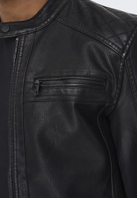 Giacca in pelle nera con design trapuntato sulle spalle, tasca verticale con cerniera sul petto e texture liscia.