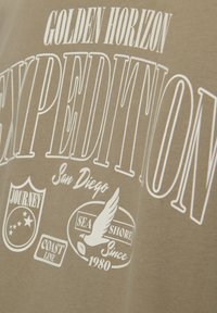 Tela beige con texto blanco "Golden Horizon Expedition", "San Diego" y logotipos que incluyen estrellas, un ala, "Sea Shore" y "Since 1980".