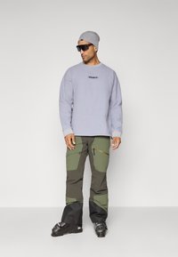 Jasnoszary sweatshirt z prążkowanymi mankietami, z logo marki; ciemnozielone spodnie cargo z kieszeniami na zamek, czarna dolna część; okulary przeciwsłoneczne i beanie.