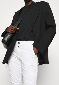 Czarna, oversize marynarka z wyraźnymi ramionami, noszona na czarnym t-shircie. Białe spodnie z czarnymi guzikami. Czarna torebka typu crossbody.
