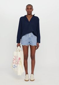 Marinblå skjorta med knappar, ljusblå denimshorts med fransade kanter, beige skor och en krämfärgad tote-bag med röda grafiska tryck.
