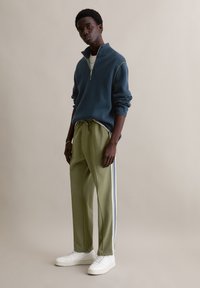 Ritsvest in marineblauw met groene stiknaden, gecombineerd met losse, olijfgroene broek met witte strepen aan de zijkant en witte sneakers.