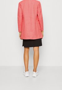 Long blazer rose texturé à coupe droite, fendue dans le dos et avec des poches latérales, assorti à une jupe noire et des baskets blanches.