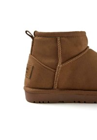 Botín de ante marrón que presenta una punta redonda, superficie texturizada, costuras decorativas y una suela de goma con relieve.