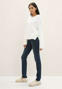 Fehér kötött pulóver laza fit-ettel, sötétkék skinny farmernadrággal és bézs vastag talpú mokaszinokkal párosítva. Egyszerű, letisztult dizájn és textúrák.