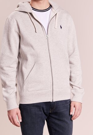 Mężczyzna w jasnoszarej bluzie na zamek z sznurkami, na białym t-shircie i ciemnych dżinsach, stojący na jednolitym tle.