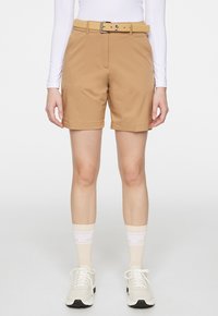 Bruna shorts i stretchmaterial med bälte och metallspänne, framfickor och avslappnad passform, i kombination med en vit långärmad skjorta.