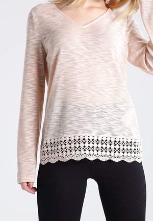 Beige long-sleeve top with subtle horizontal pattern and scalloped lace trim at hem, worn with black fitted pants.