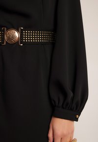 Czarna sukienka z teksturowanym, szerokim czarnym paskiem z złotymi ćwiekami i okrągłą, metalową klamrą. Długie, bufiaste rękawy z guzikiem przy mankiecie.