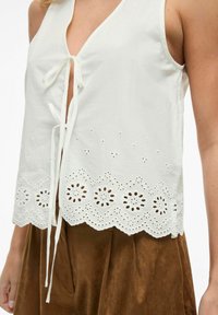 Hvit ermleløs topp med v-hals og knytelukking. Har øyelet broderi langs falden med blomsterdesign. Kombinert med brune bukser.