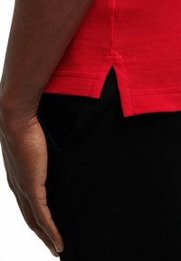 Polo shirt rossa con spacchi laterali, abbinata a pantaloni neri di tessuto strutturato. Il tessuto sembra resistente; la camicia ha maniche corte e un taglio classico.