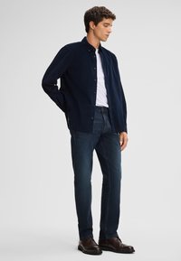 Marineblaue Knopfleiste-Hemd aus strukturiertem Stoff, über einem weißen T-Shirt getragen, kombiniert mit dunkelblauen Jeans und braunen Schuhen, Frontalansicht.