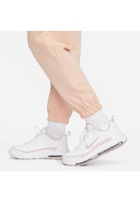 Baltie Nike sporta apavi ar rozā akcentiem, ar teksturētu sietu, mīkstu zoli un redzamu gaisa amortizāciju. Kombinēti ar rozā sporta biksēm.