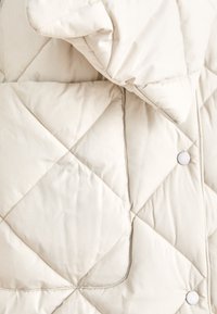 Casaco bege acolchoado com colarinho alto, padrão de diamante e botões de pressão brancos. Textura suave e lisa com uma aparência almofadada.