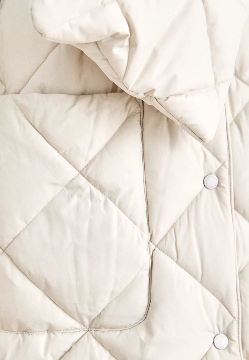 Beige gesteppte Jacke mit hohem Kragen, Rautenmuster und weißen Druckknöpfen. Weiche, glatte Textur mit gepolsterter Optik.