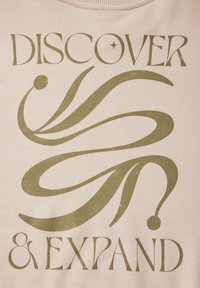 Una tela de algodón beige presenta un texto verde intenso que dice "DESCUBRE Y EXPANDE", acompañado de espirales decorativas y un acento circular.