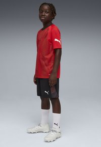 Rød kortærmet trøje med bølgemønster, sorte shorts med røde detaljer, hvide strømper og sølvfarvede fodboldstøvler. Puma-logo på trøje, shorts og sko.
