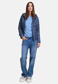 Marineblauwe pufferjack met horizontale stiksels, gecombineerd met een lichtblauwe trui en losse blauwe jeans. De schoenen zijn chunky en reflecterend wit.
