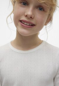 Vit långärmad skjorta med en subtil strukturerad mönster och vågig halsringning, gjord av lätt tyg, bärs av ett barn med ljust hår.