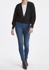 Cárdigan negro con un profundo escote en V y dos botones, combinado con jeans ajustados azules y botines. Textura suave, diseño ajustado.