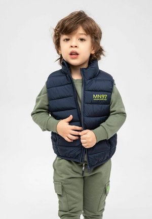 Gilet matelassé bleu marine avec motif de coutures, logo jaune sur la poitrine, par-dessus une chemise à manches longues vertes et un pantalon de jogging vert assorti avec poches.