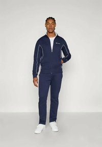 Tengerészkék szabadidőruha fehér részletekkel. A cipzáras dzsekin álló gallér található; a nadrág fitten illeszkedik. Fehér sportcipővel viselve.