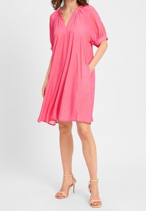 Woman wearing loose, knee-length pink dress with subtle leopard print, short sleeves, and beige high-heeled sandals, standing with one hand in pocket.