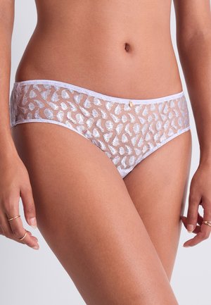 Femme portant un sous-vêtement hipster en dentelle blanche transparente imprimé léopard, montrant le ventre et le haut des cuisses, les mains relâchées le long du corps.