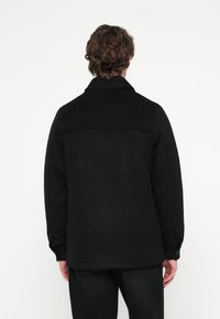 Sort jakke lavet af tekstureret stof med krave, bagstykke og lange ærmer. Kanten har afrundede kanter og manchetter med knapper.